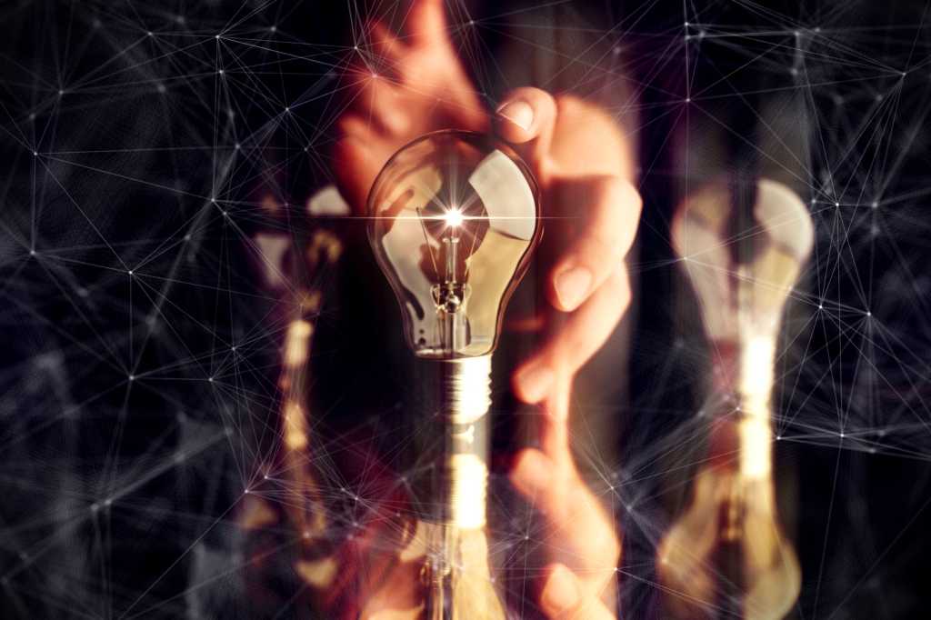 A hand picks a glowing lightbulb with abstract technology overlay. / ideas / innovation / creativity