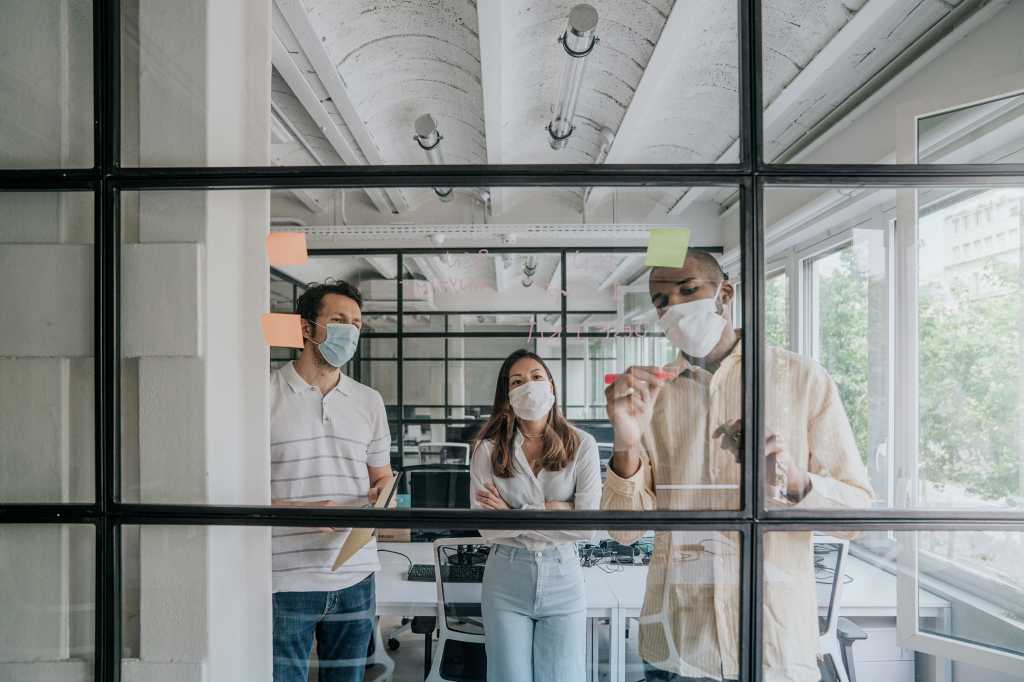Co-workers wear face masks while working at a dry-erase glass wall in a post-COVID office workspace.
