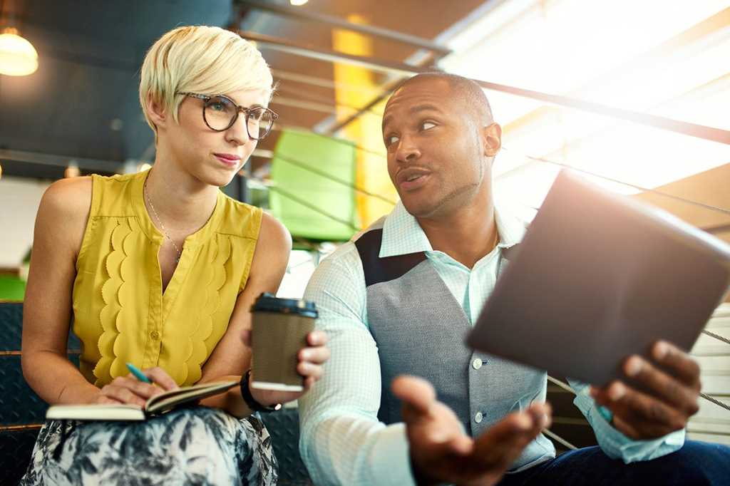 A woman and a man have a conversation while holding a notebook and pen, coffee and a mobile tablet.
