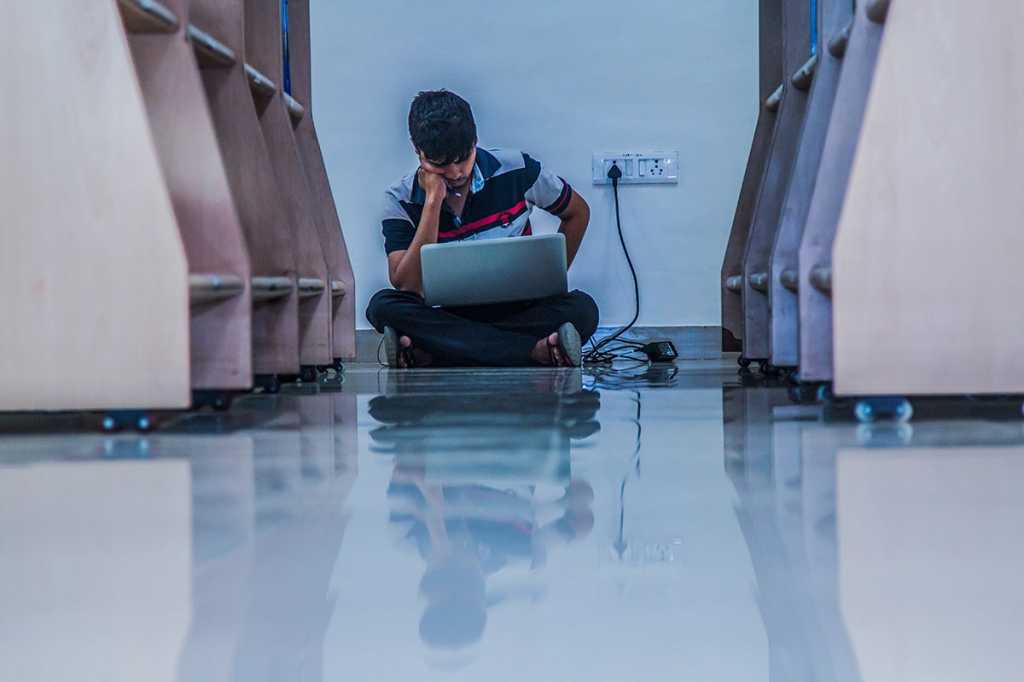 A solitary man sits between shelves near an outlet while charging his laptop.