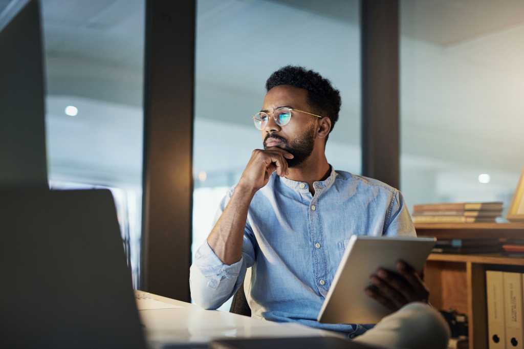 Man at desk with ipad, looking at computer screen thinking