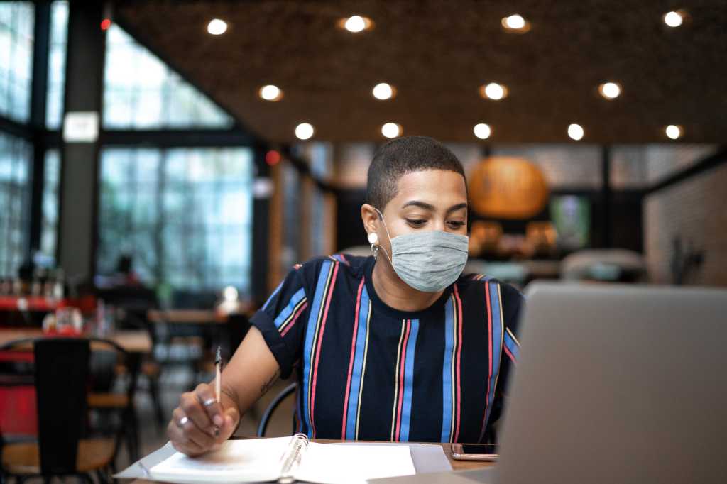 A laptop user wearing a protective face mask takes notes in an open workspace.