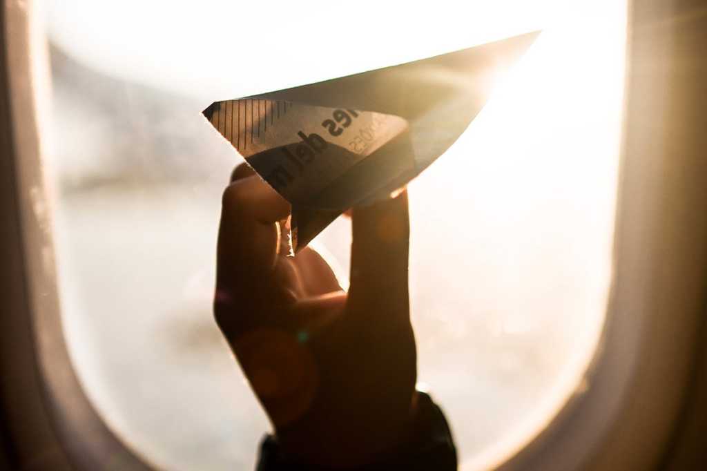 A passenger holds a paper airplane up to a window in flight. / travel / journey / transportation