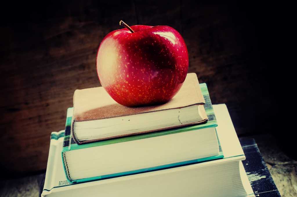 A stack of school books sits on a desk, with an apple on top. [Education/Learning]