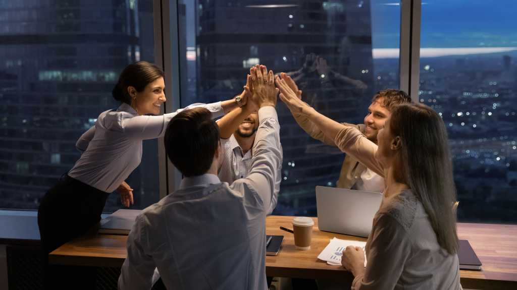 Motivated multinational team raise high fives on briefing after finding problem solution as successful brainstorm result. Happy workers unite hands above conference desk celebrate common achievement