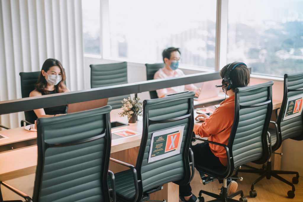 Co-workers maintain social distancing as signs mark chairs to remain vacant in a post-COVID office.