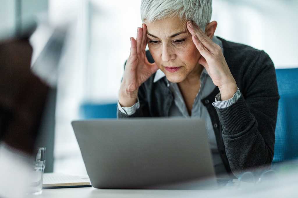 A miserable, stressed laptop user holds her head in her hands.
