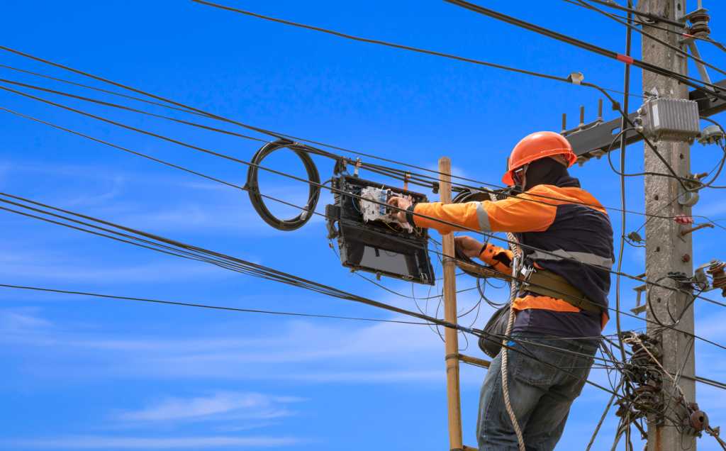 Technician on wooden ladder is installing fiber optic system in internet splitter box on electric pole against blue sky