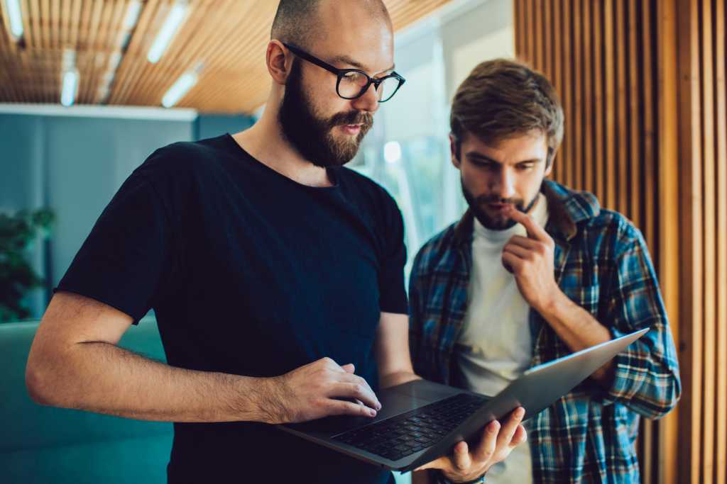 Concentrated bearded male in eyeglasses standing with marketing team member in cafe with laptop and looking at screen together while working on project document for startup