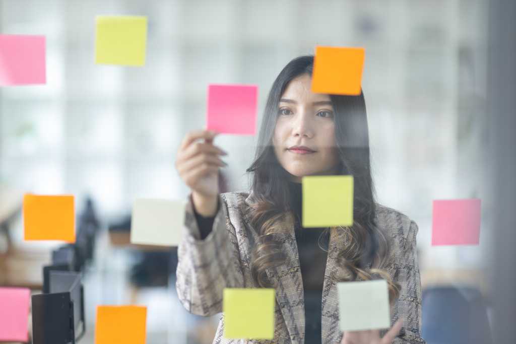 Business female employee with many conflicting priorities arranging sticky notes commenting and brainstorming on work priorities colleague in a modern office.
