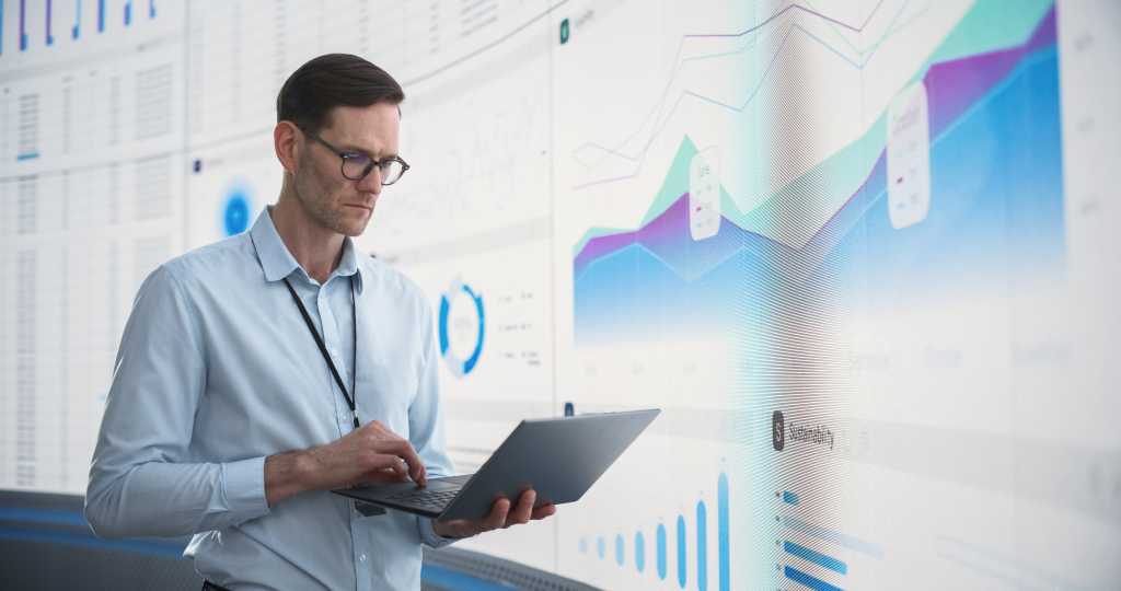 Caucasian Male Business Analyst With Laptop Computer Standing Next To Big Digital Screen With Graphs And Charts. Successful Man Assessing Risks And Developing Strategies For Company Growth, Expansion.