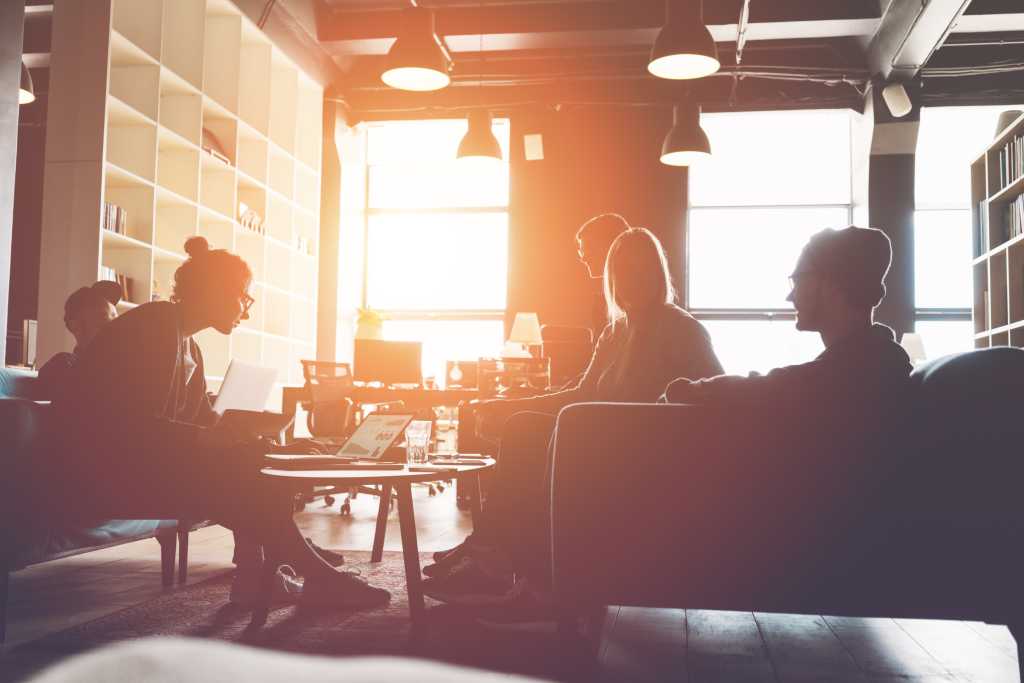 Silhouette of business group brainstorming new plan. Team meeting on the couch. Big open space office. Five people. Intentional sun glare