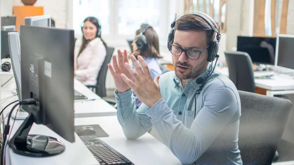 junger gut aussehender männlicher technischer Support-Mitarbeiter, der versucht, einem Kunden etwas zu erklären, während er im Callcenter mit einem Freisprech-Headset
