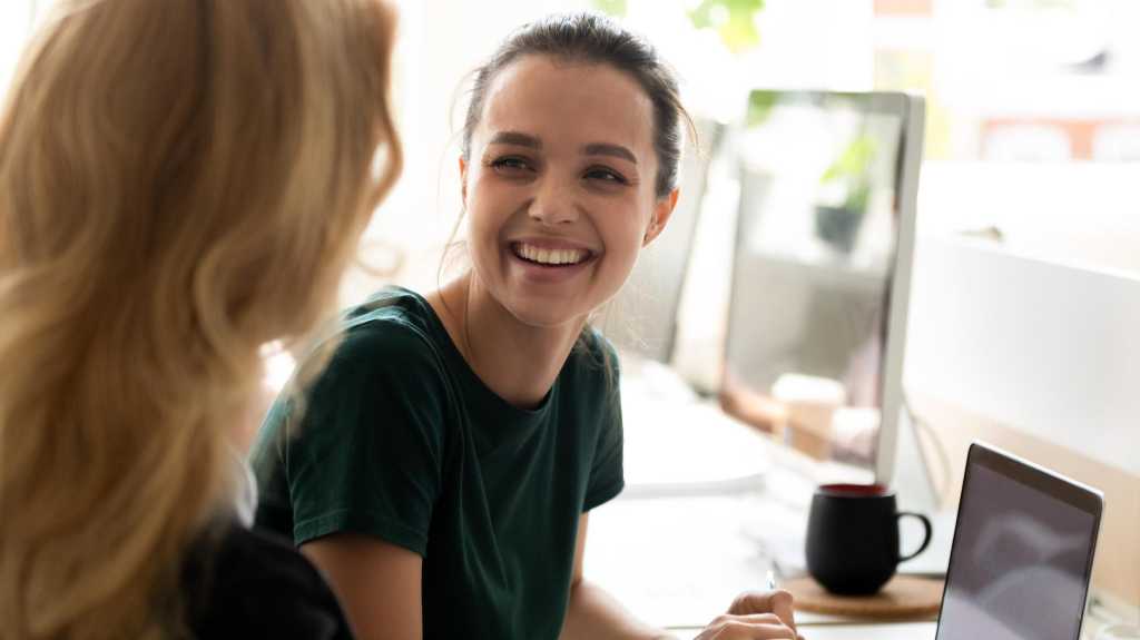 Happy millennial intern, student girl talking to teacher, consulting tutor, mentor at computer in training center, discussing new professional skills, smiling, laughing. Business education, internship