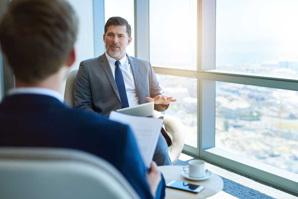 Handsome mature corporate manager sitting in a modern office space, holding a digital tablet while interviewing a young business applicant