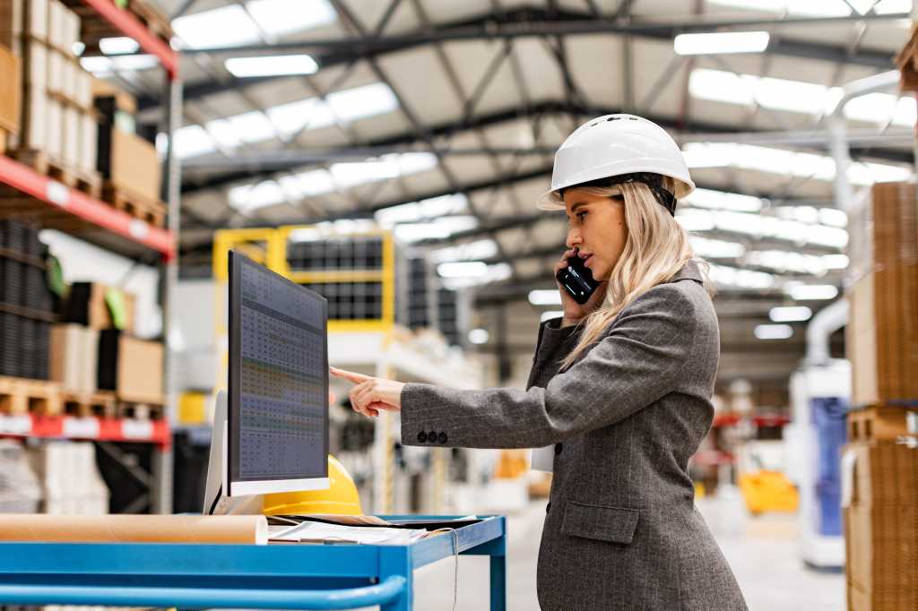 Female engineer, project manager standing in modern industrial factory by computer, making phone call. Team management in manufacturing facility.