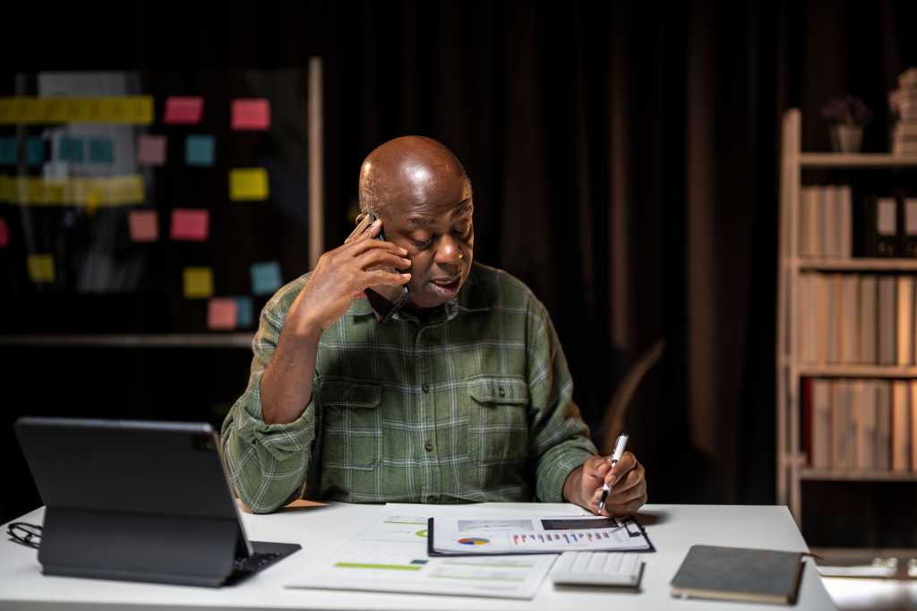 A man is sitting at a desk with a laptop and a piece of paper. He is looking at the paper and writing on it. The scene suggests that he is working on a project or task