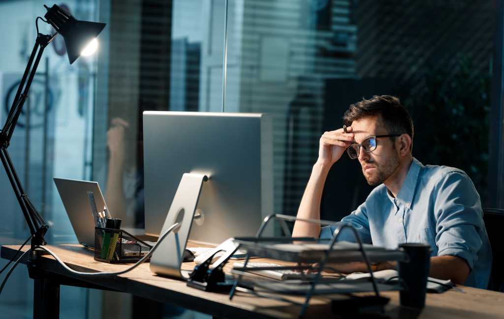 Casual tired office worker sitting at desk using computer and doing overtime project in lamplight.