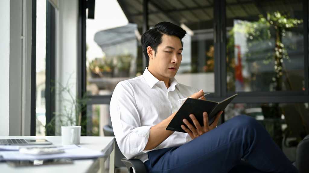 Professional and handsome millennial Asian businessman or male boss focuses on reading a book or checking his meeting schedule on his planner book while sitting at his desk.