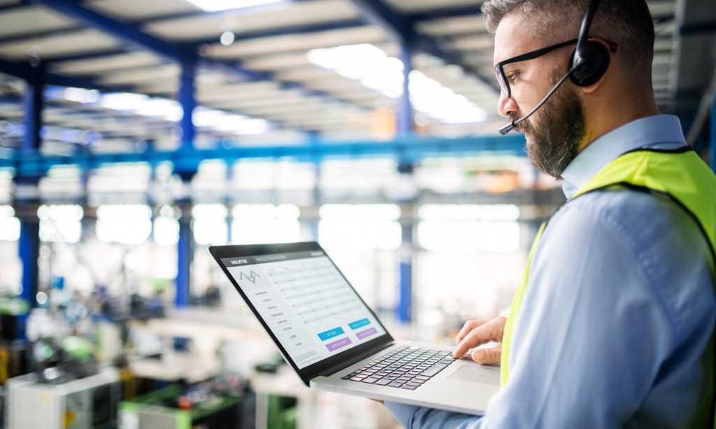 Side view of technician or engineer with headset and laptop standing in industrial factory.