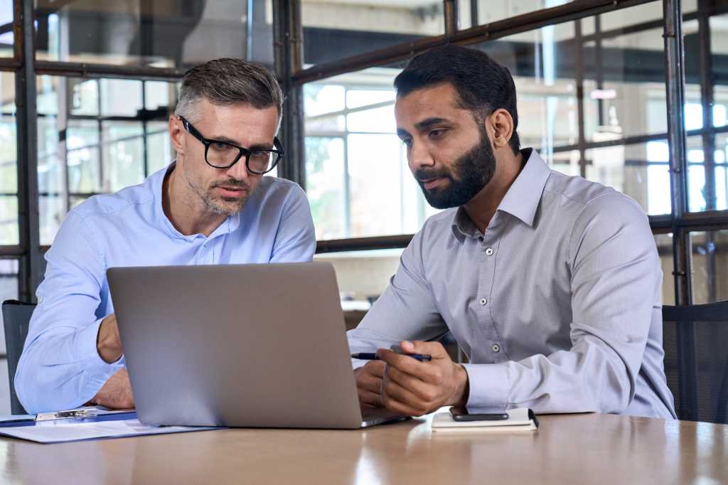 Two diverse business men analysts partners talking, working together, discussing data management using laptop computer. Indian manager consulting client with online technology at office meeting.