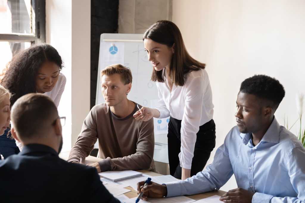 Young female employee sharing point of view with focused diverse teammates at brainstorming meeting. Group of international employees discussing project ideas, financial report, marketing strategy.