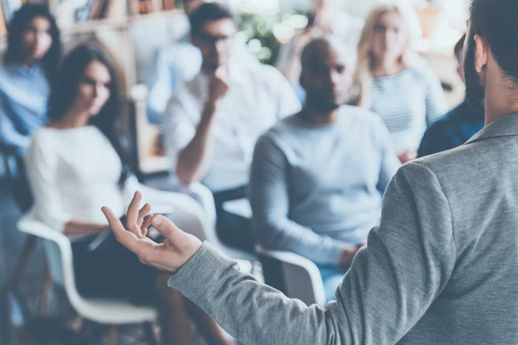 Business coach. Rear view of man gesturing with hand while standing against defocused group of people sitting at the chairs in front of him
