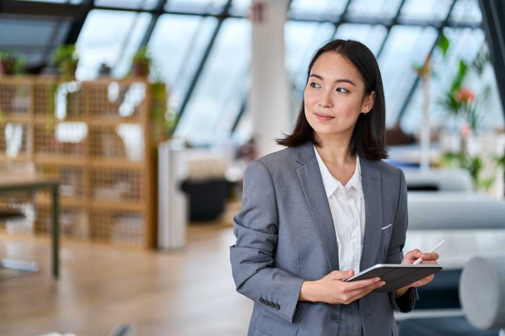 Young Asian business woman entrepreneur standing in office holding digital tablet. Businesswoman leader, professional company manager using smart corporate management technology looking at copy