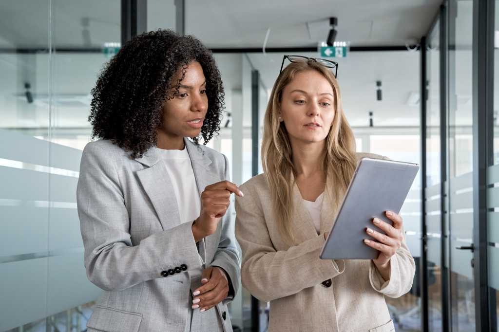 Two businesswoman working together using digital tablet pc and talking about a business project. Small creative diversity team of African American and blond females executives meeting work in office.