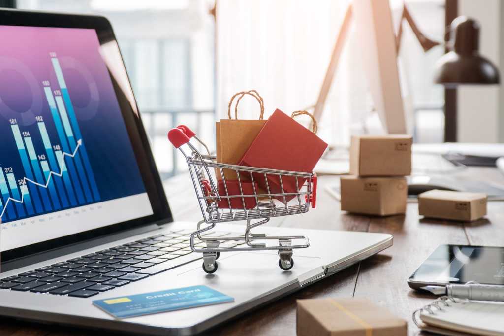 A miniature shopping cart with shopping bags, cardboard boxes sitson a laptop with a credit card.