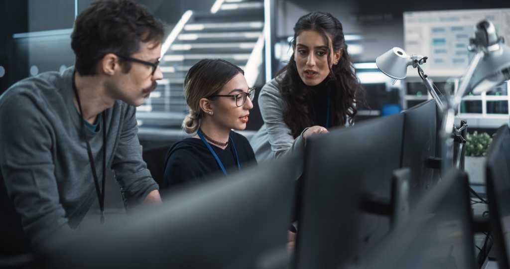 Team of Three Talented Young Software Engineers Use Computer to Discuss a Technological Project in Modern Industrial Office. Group of Male and Female Scientists Work in Research and Development Center