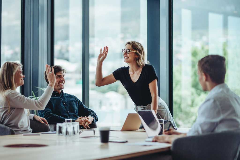Businesswoman giving a high five to her coworker while their male colleagues sitting by. Coworkers sitting in meeting room celebrating project success.