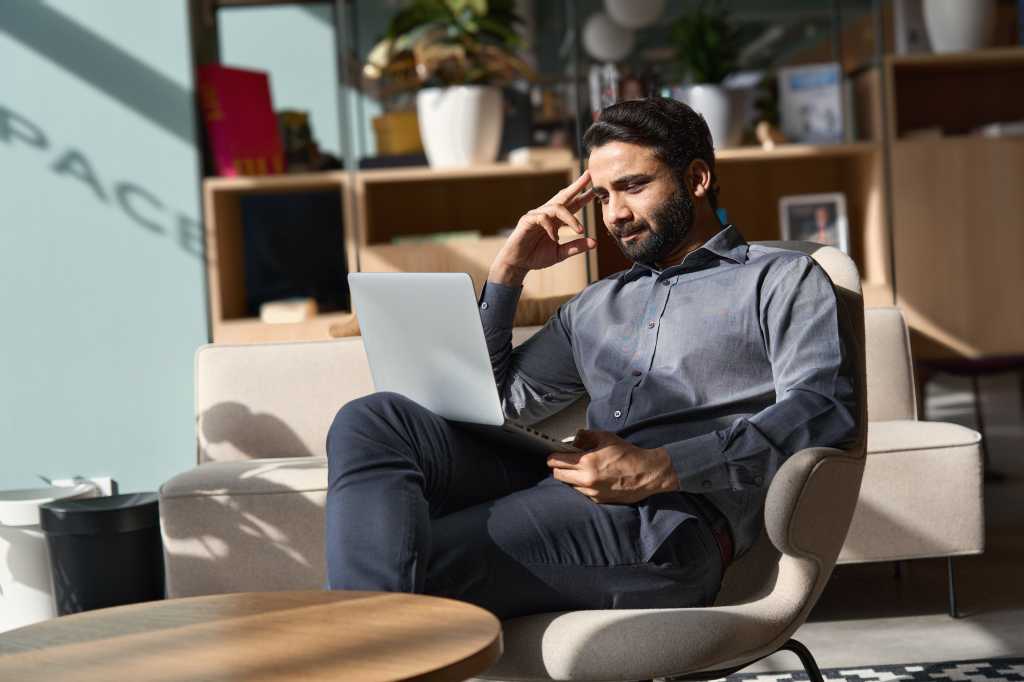 Indian business man executive working on laptop sitting on chair in modern office. Eastern male entrepreneur using computer remote studying, virtual training, watching online education webinar.