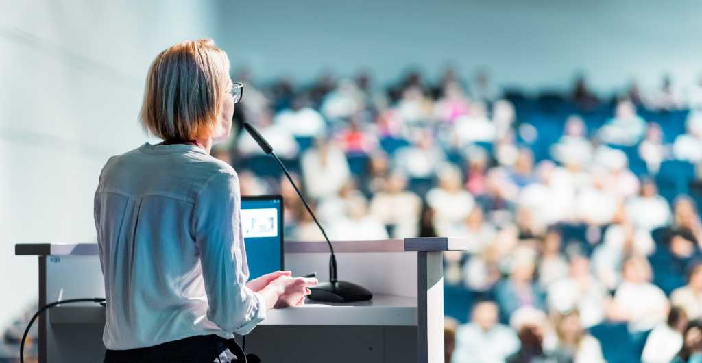 Female speaker giving a talk on corporate business conference. Unrecognizable people in audience at conference hall. Business and Entrepreneurship event