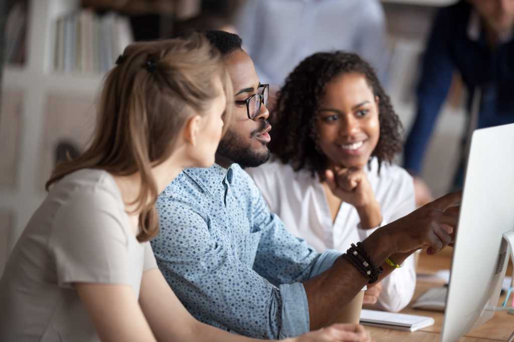 Diverse businesspeople working in shared office, focus on black american team leader sitting at desk together with females colleagues, mentor helps understand research data company corporate program