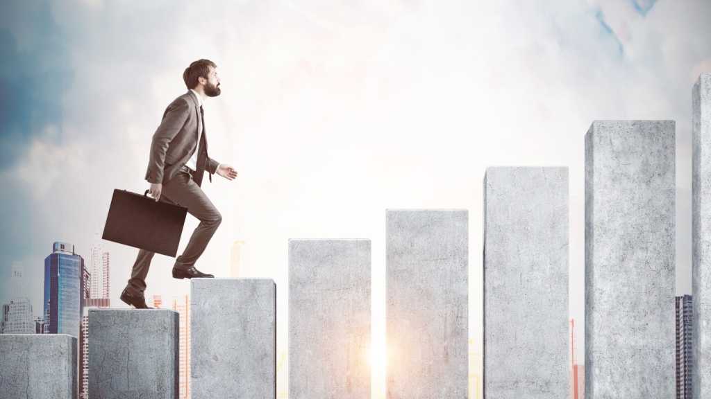 A man in a suit holding a briefcase is walking up a series of cement pillars. A cityscape can be seen in the background.