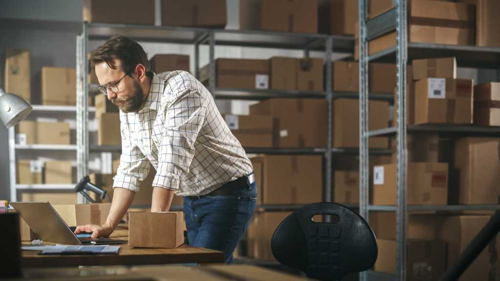 Inventory Manager Preparing a Small Cardboard Parcel for Postage. Stylish Young Male Small Business Owner Working on Laptop Computer in Warehouse Facility