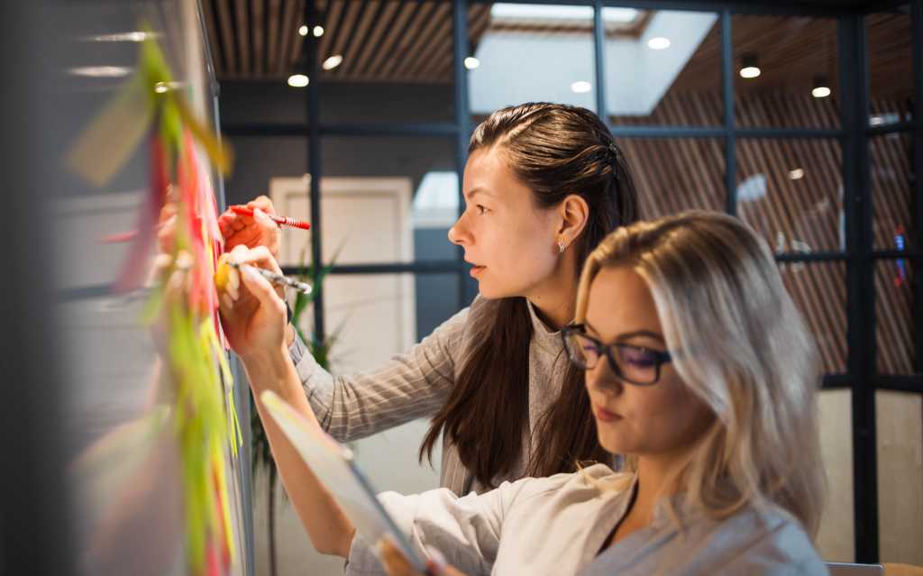 Teamwork, project management, agile methodology. Two young business women in the office are planning product development and support. Colleagues glue sticky stickers on the Board