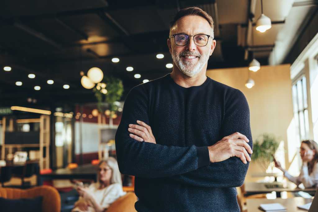 Mature businessman standing in a co-working space. Happy mature businessman smiling at the camera while standing with his arms crossed. Experienced entrepreneur working in a modern workspace.