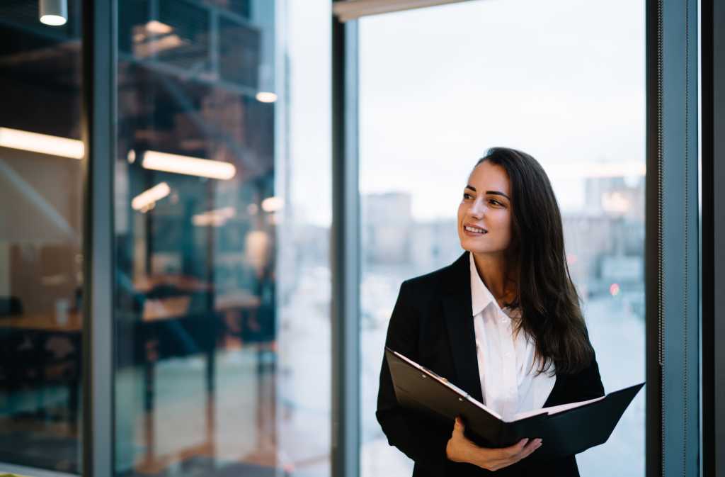 Young smiling female entrepreneur in formal wear standing with documents near panoramic window during work in contemporary company looking away