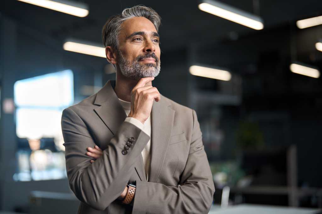 Middle aged business man investor, doubtful mature male ceo executive wearing suit standing in office holding hand in chin looking away at work thinking on leadership question, feeling doubt.