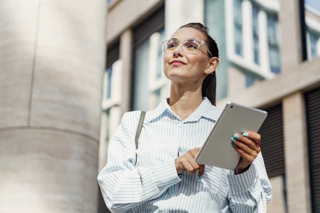 A visionary businesswoman holds her tablet with poise, gazing forward optimistically against a backdrop of modern urban architecture.