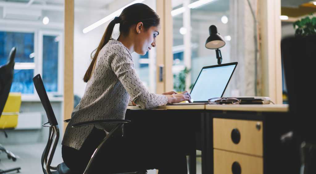 Side view of focused woman in casual clothes with dark hair sitting at desk and typing on netbook in modern workspace