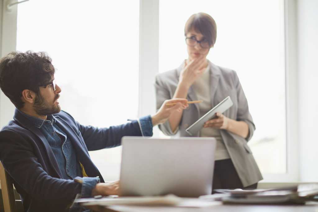 Young successful middle eastern man looking up from laptop to consult young colleague standing by him with tablet, pointing at it