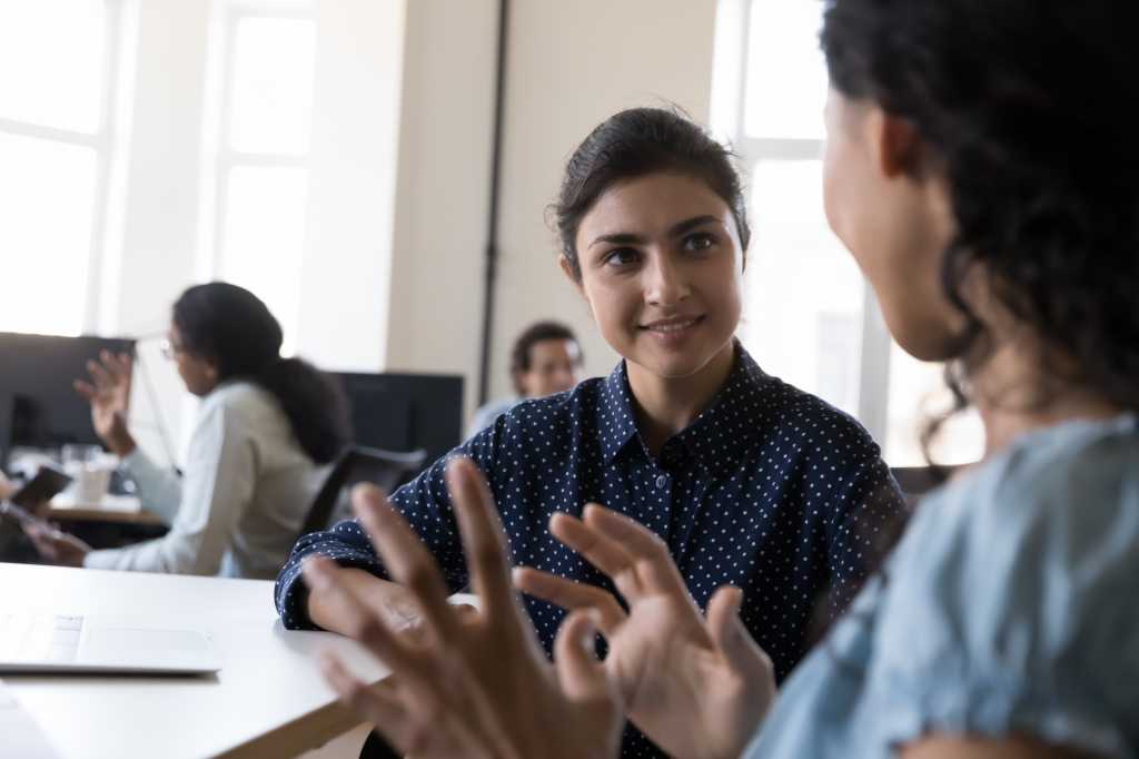 Happy Indian intern listening to mentor, consulting corporate teacher at shared office workplace in training center, discussing new professional skills, smiling. Business education, internship