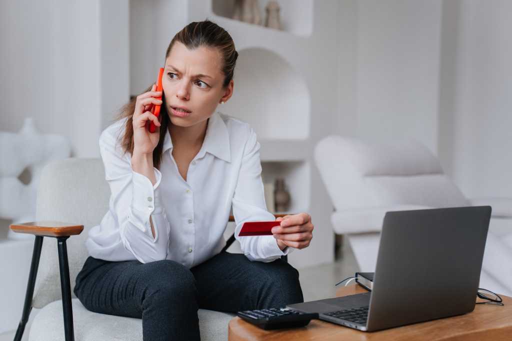 Worried businesswoman talks by phone with bank employee holds credit card sitting at desk with laptop tries to resolve problems, irritated by partners mistakes. Crisis, failure.