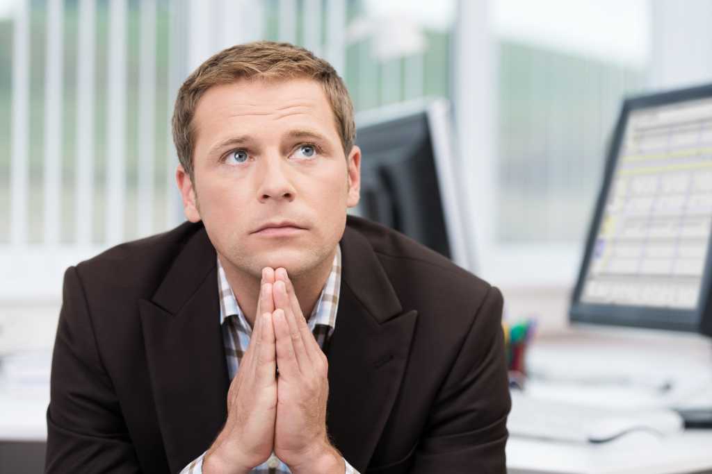 Worried pensive businessman sitting at his desk staring off into space with his hands folded under his chin