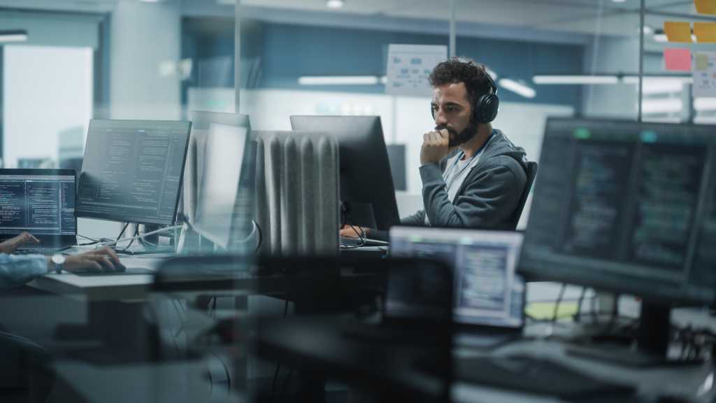 Diverse Office: White IT Programmer Working on Computer. Male Specialist Creating Innovative Software. Engineer Developing App, Program, Video Game. Wearing Headphones Listening Podcast, Music