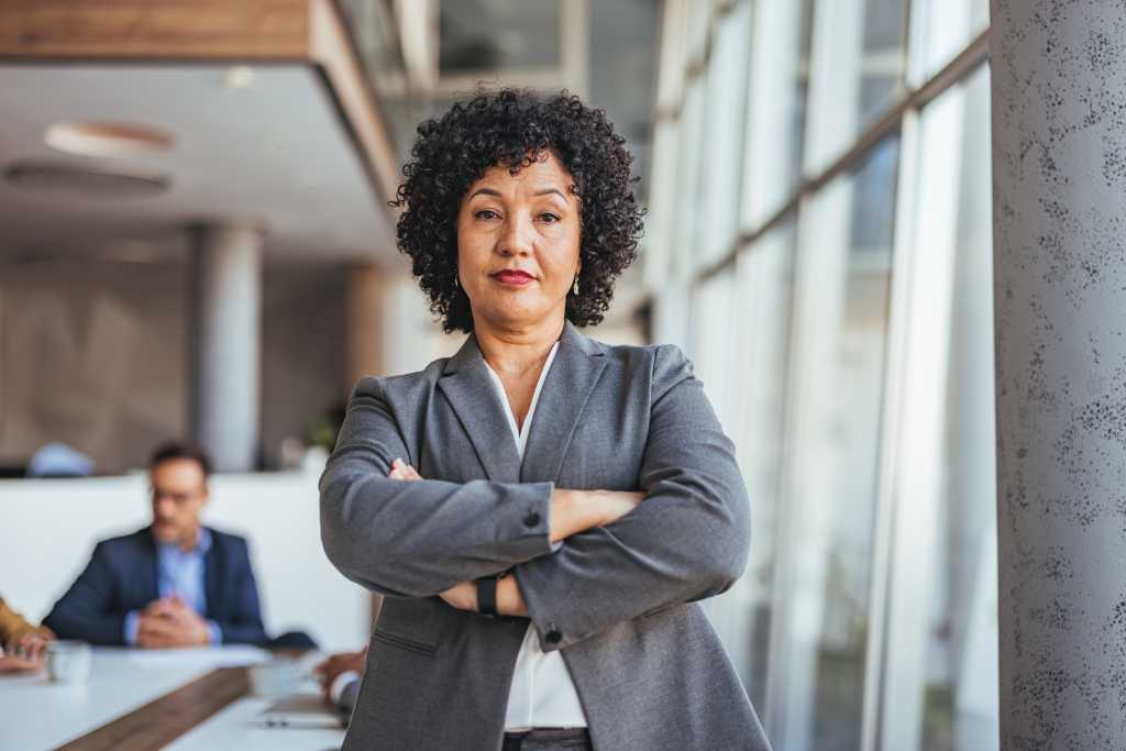 A confident businesswoman with crossed arms stands in a modern office, exuding leadership and professionalism. Natural light from large windows enhances the dynamic workplace setting.