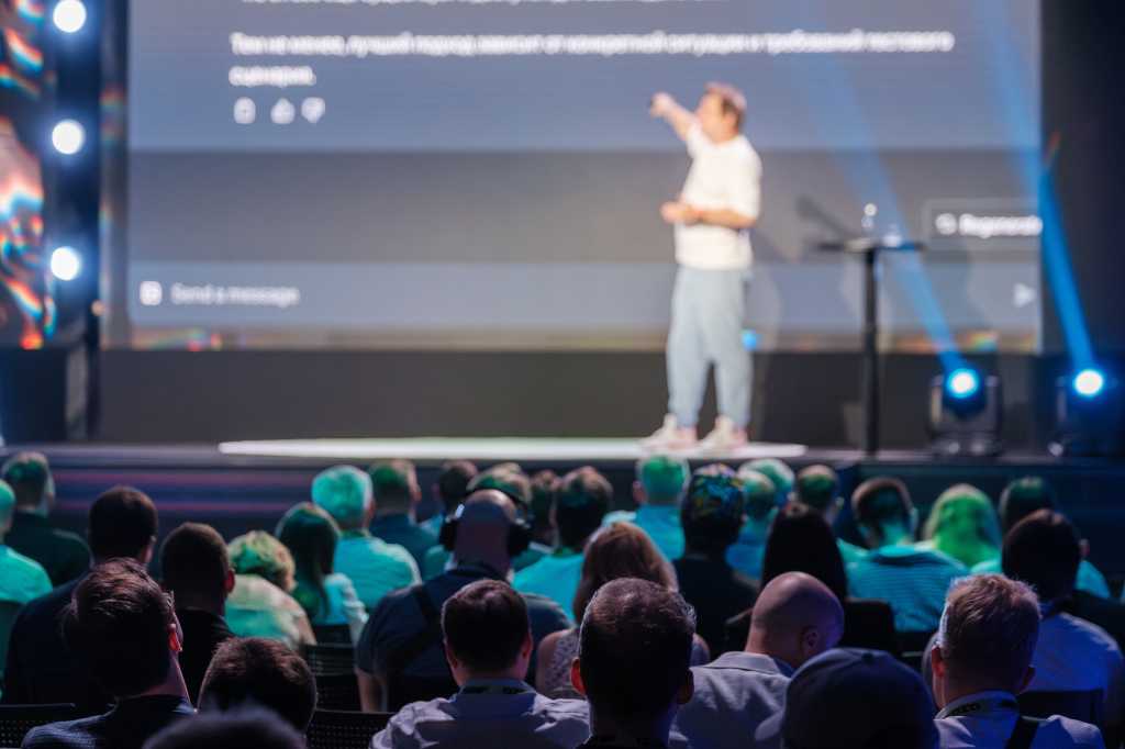 Business professional presenting at a conference with a large audience, highlighting key points on a screen.